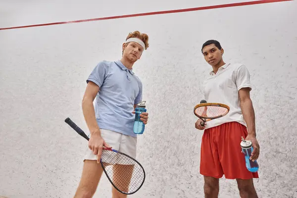 Jovens desportistas inter-raciais com raquetes de squash e garrafas com água em pé dentro do tribunal — Fotografia de Stock