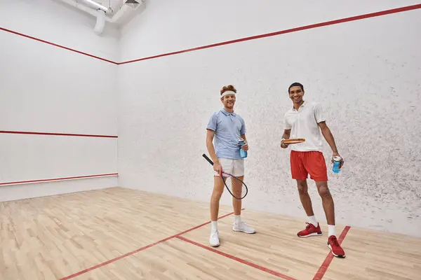 Cheerful interracial sportsmen with squash racquets and bottles with water standing inside of court — Stock Photo