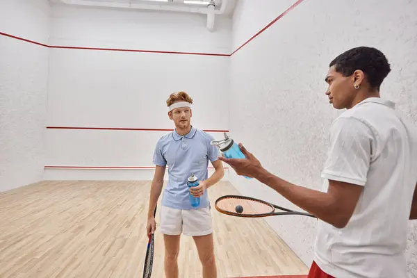Esportistas inter-raciais com raquetes de squash e garrafas com água em pé dentro do tribunal — Fotografia de Stock