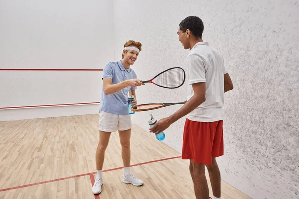 Heureux sportifs interracial avec des raquettes de squash et des bouteilles avec de l'eau debout dans la cour — Photo de stock