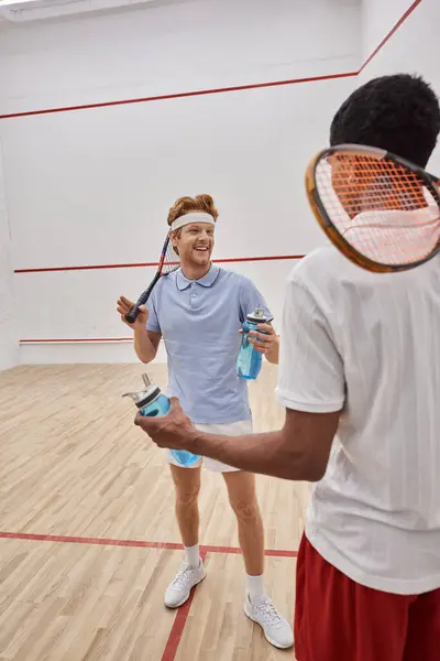 Smiling interracial sportsmen with squash racquets and bottles with water chatting  in court — Stock Photo