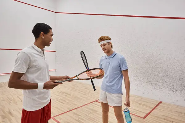 Joyful interracial male friends with squash racquets and bottles with water chatting  in court — Stock Photo