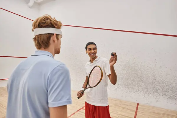 Happy african american man with squash ball and racquet standing near friend inside of court — Stock Photo