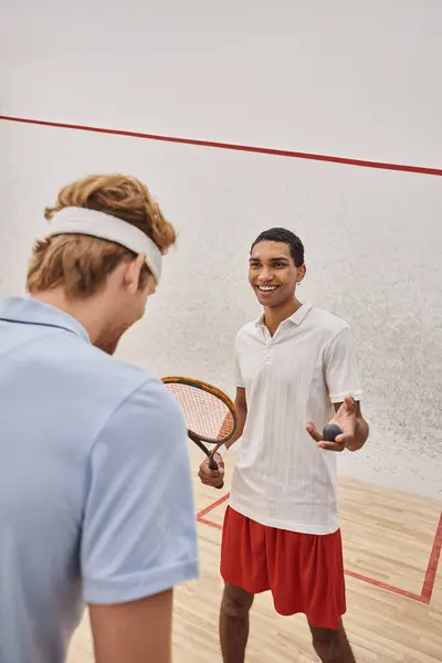 Cheerful african american man with squash ball and racquet standing near friend inside of court — Stock Photo