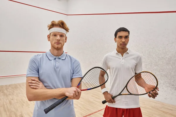 Зосереджені міжрасові друзі чоловічої статі в спортивному одязі, що стоять разом із сквош-ракетками в суді — стокове фото