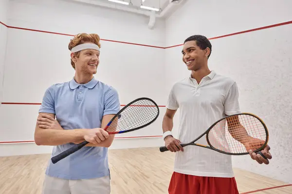 Happy interracial male friends in sportswear standing together with squash racquets in court — Stock Photo