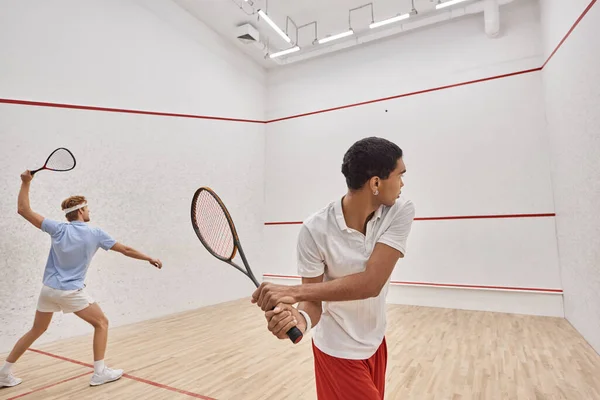 Amis interraciaux en vêtements de sport jouer au squash ensemble à l'intérieur de la cour, mode de vie actif — Photo de stock