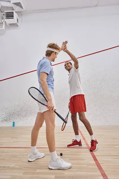 Cheerful multicultural friends in sportswear giving high five after playing squash in court — Stock Photo