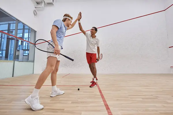 Dos amigos multiculturales felices en ropa deportiva dando cinco altos después de jugar squash en la corte - foto de stock