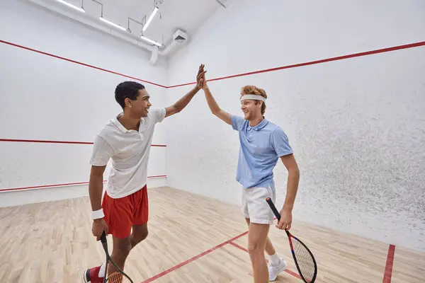 Smiling multicultural friends in sportswear giving high five after playing squash in court — Stock Photo