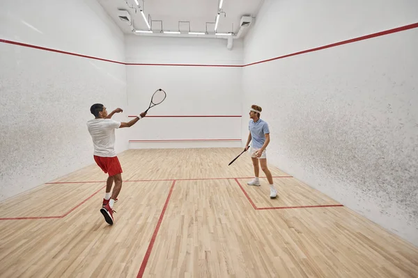 Hombres multiculturales en ropa deportiva jugando squash juntos dentro de la cancha, motivación y deporte - foto de stock
