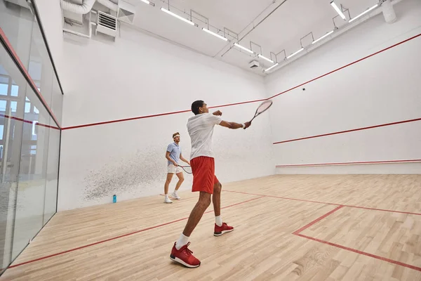 Hombres atléticos multiculturales en ropa deportiva jugando squash juntos dentro de la corte, motivación - foto de stock