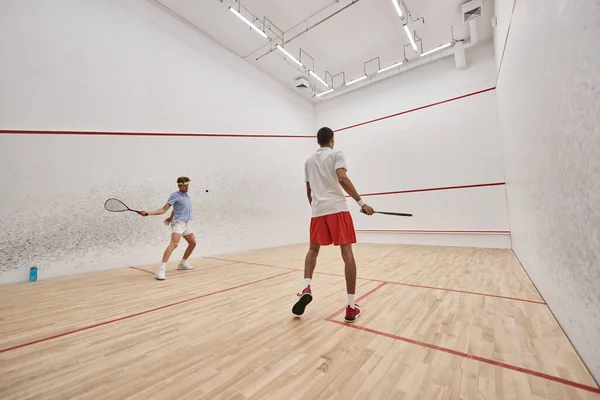 Hombres interraciales y atléticos en ropa deportiva jugando squash juntos dentro de la corte, motivación - foto de stock
