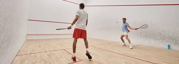 Hommes sportifs interracial en vêtements de sport jouant au squash ensemble à l'intérieur du court, bannière de motivation — Photo de stock