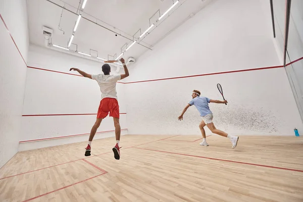 Young and active interracial sportsmen in sportswear jumping while playing squash inside of court — Stock Photo
