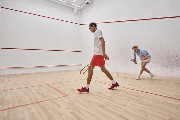 Hombres activos y diversos en ropa deportiva jugando squash dentro de la cancha, desafío y motivación - foto de stock
