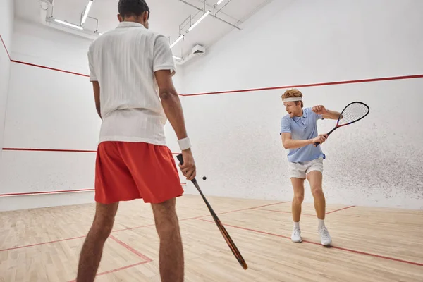 Hombres multiétnicos activos en ropa deportiva jugando squash dentro de la corte, desafío y motivación - foto de stock