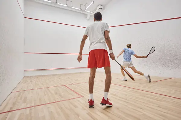 Interrazziale uomini attivi in abbigliamento sportivo giocare a squash all'interno della corte, sfida e motivazione — Foto stock