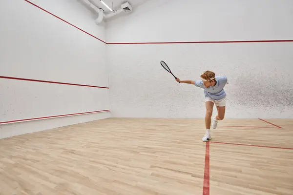 Joven y activo pelirrojo en ropa deportiva jugando squash dentro de la cancha, desafío y motivación - foto de stock