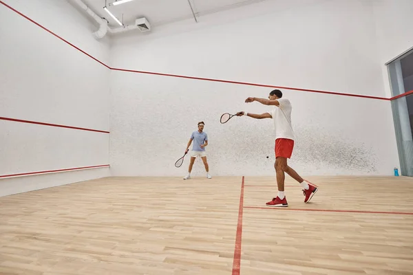 Jugadores multiétnicos activos en ropa deportiva jugando squash dentro de la cancha, esfuerzo y motivación - foto de stock