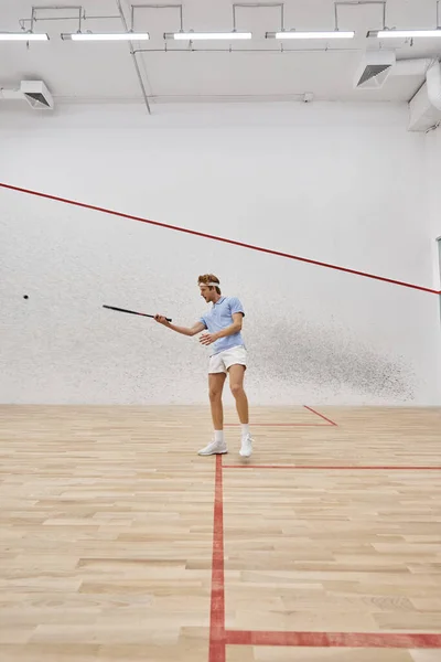 Young redhead sportsman in headband playing squash inside of court, challenge and motivation — Stock Photo