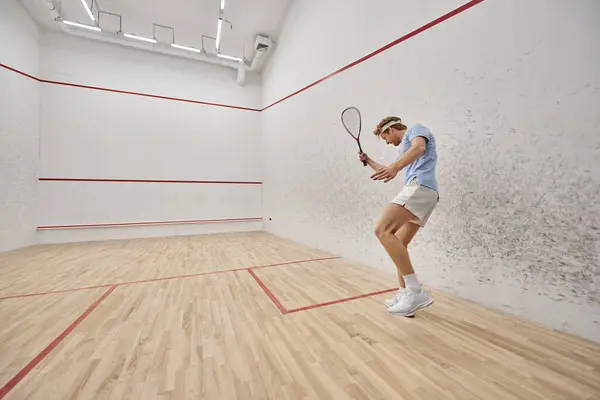 Young and redhead sportsman in headband playing squash inside of court, challenge and motivation — Stock Photo