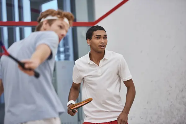 Emocionado hombre afroamericano sosteniendo raqueta de squash mientras juega con un amigo en primer plano borroso - foto de stock