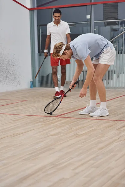Ruiva homem na cabeça dobra sobre squash bola no chão dentro da quadra, interracial amigos — Fotografia de Stock
