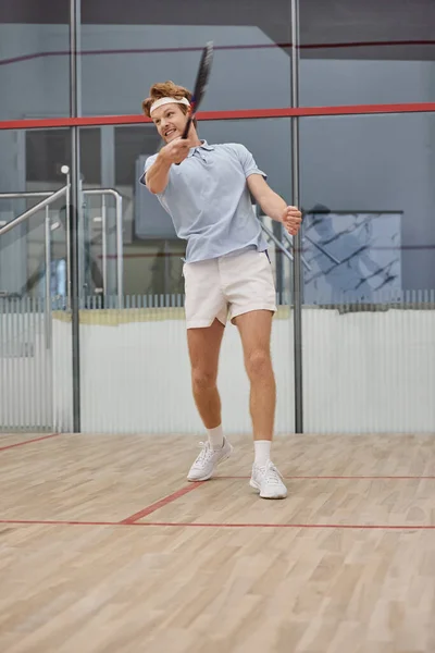 Heureux rousse homme dans bandeau flexion sur balle de squash sur le sol à l'intérieur du court, amis interracial — Photo de stock
