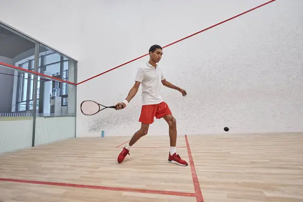 Esportista afro-americano em shorts esportivos vermelhos segurando raquete enquanto joga squash dentro da corte — Fotografia de Stock