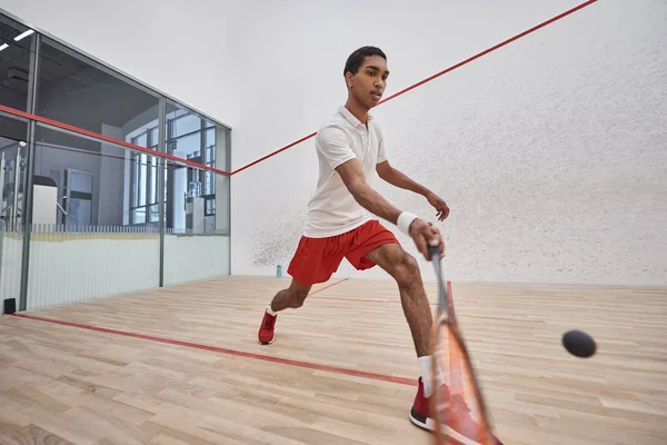 Sportif afro-américain en short de sport rouge tenant la raquette tout en jouant au squash, motion shot — Photo de stock