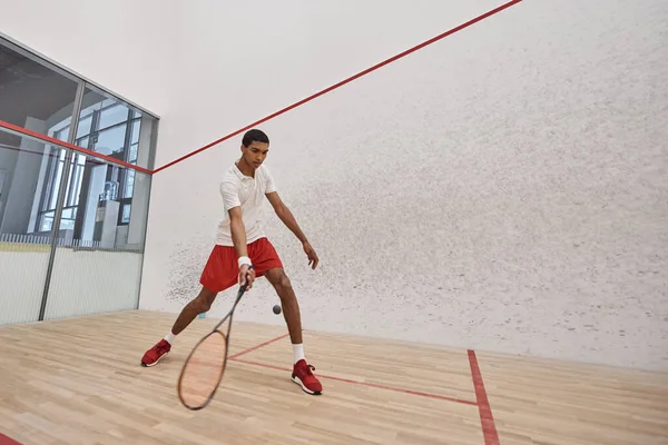 Jovem afro-americano em shorts esportivos vermelhos segurando raquete enquanto joga squash, tiro movimento — Fotografia de Stock