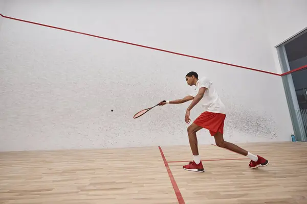 Deportista afroamericano activo en pantalones cortos rojos sosteniendo raqueta mientras juega squash dentro de la corte - foto de stock