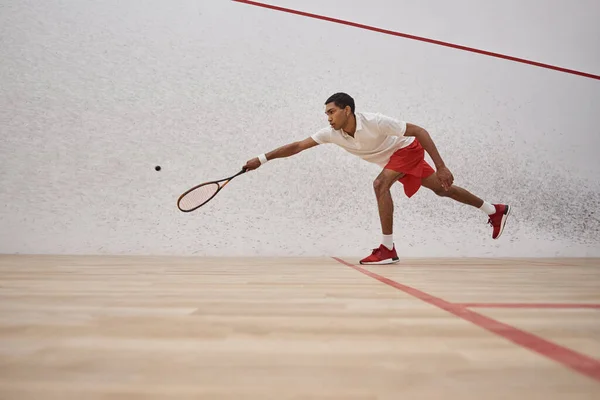 Sportif afro-américain en short rouge tenant raquette et jouant au squash à l'intérieur du court, sport — Photo de stock