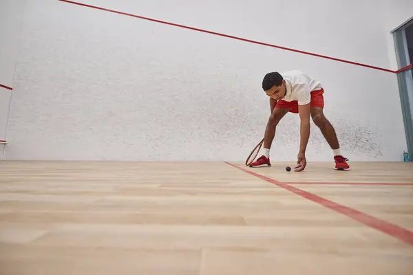 Homme afro-américain en short rouge tenant raquette et ramasser balle de squash à l'intérieur du court — Photo de stock