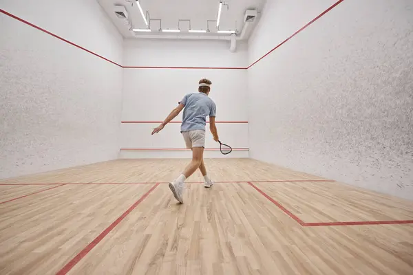 Atlético y pelirrojo deportista sosteniendo raqueta mientras juega squash dentro de la cancha, tiro de movimiento - foto de stock