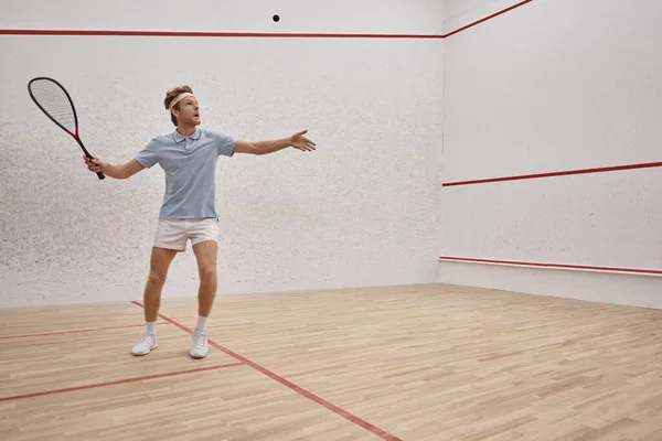 Atlético pelirrojo hombre en ropa deportiva celebración de raqueta y jugar squash dentro de la cancha, tiro de movimiento — Stock Photo