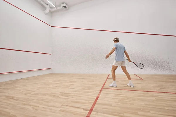Deportivo y pelirrojo hombre sosteniendo raqueta mientras juega squash dentro de la cancha, tiro de movimiento - foto de stock