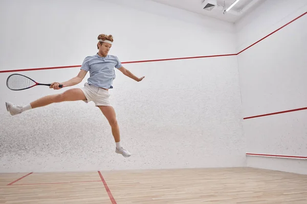 Tiro en movimiento, divertido deportista pelirrojo sosteniendo raqueta y saltando mientras juega squash en la corte - foto de stock