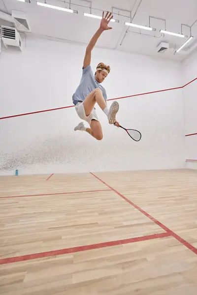 Movimento e diversão, esportista ativo segurando raquete e pulando enquanto joga squash dentro do tribunal — Fotografia de Stock
