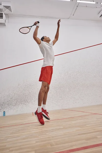 Movimiento, enérgico hombre afroamericano sosteniendo raqueta mientras salta y juega squash, entrenamiento - foto de stock