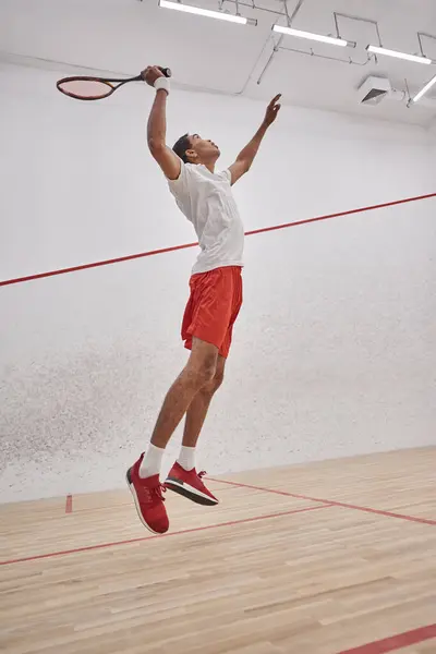 Foto en movimiento, enérgico deportista afroamericano sosteniendo raqueta mientras salta y juega squash - foto de stock
