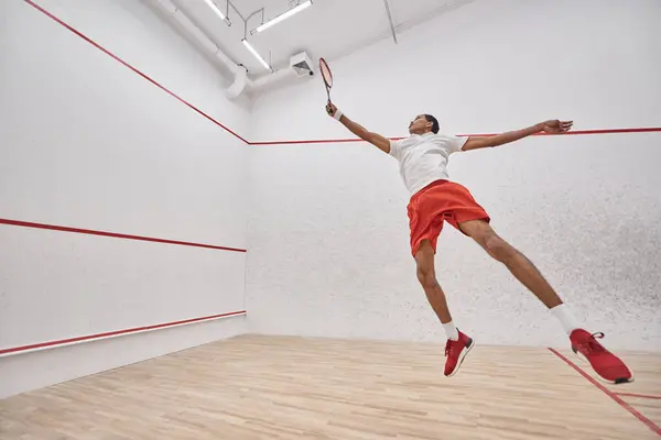 Movimiento, enérgico deportista afroamericano con raqueta saltando y jugando squash en la corte - foto de stock