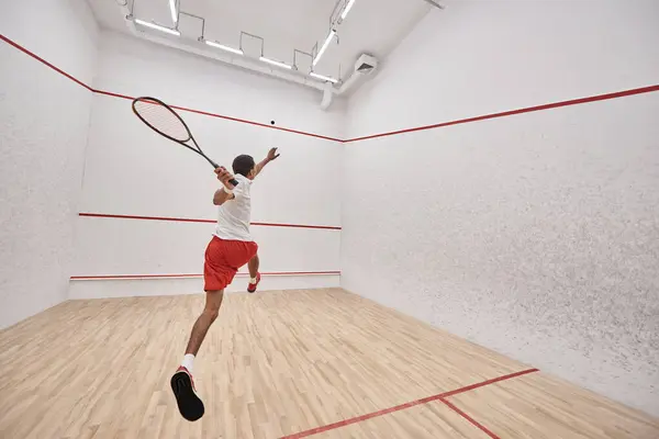 Jovem atlético afro-americano jogador com raquete pulando e jogando squash na corte, visão traseira — Fotografia de Stock
