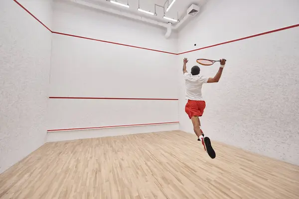 Atlético afro-americano jogador com raquete pulando e jogando squash na corte, visão traseira — Fotografia de Stock