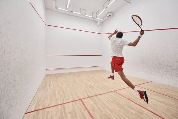 Atletico giocatore afroamericano con racchetta che salta mentre gioca a squash in tribunale, vista posteriore — Foto stock