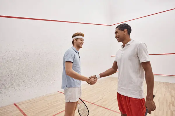 Happy interracial sportsmen in active wear smiling and shaking hands while holding squash racquets — Stock Photo