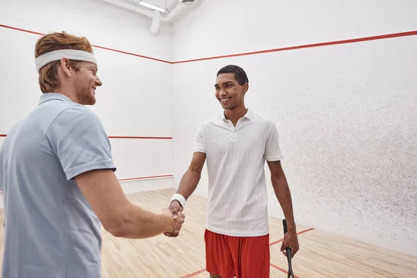 Fröhliche interrassische Sportler in aktiven Gewändern lächeln und schütteln Hände, während sie Squash-Schläger in der Hand halten — Stockfoto