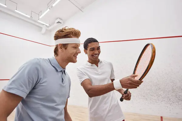 Feliz interracial desportistas no ativo desgaste sorrindo e verificando squash raquete dentro do tribunal — Fotografia de Stock