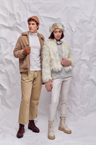 Asian woman in faux fur jacket and mittens looking at camera near trendy man on white paper backdrop — Stock Photo